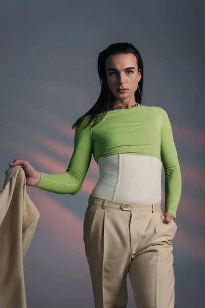 Brunette queer model in corset holding jacket and looking at camera on abstract background — Stock Photo