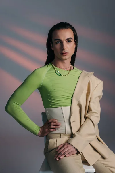 Trendy young queer model in suit and corset posing on chair on abstract background — Foto stock