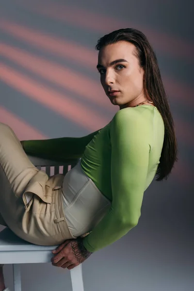 Trendy brunette nonbinary model sitting on chair and looking at camera on abstract background — Stock Photo