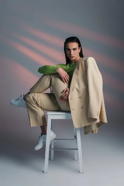 Брюнетка небинарная модель в стильном бежевом костюме сидит на стуле на абстрактном фоне — стоковое фото