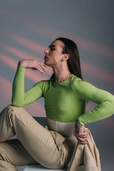 Side view of trendy queer model holding jacket while sitting on chair on abstract background — Stock Photo