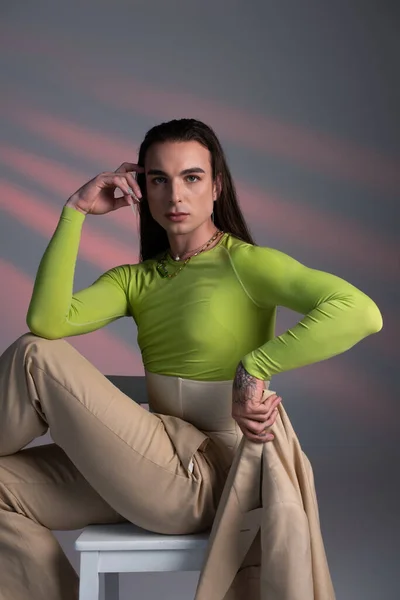 Young brunette queer person holding jacket while sitting on chair on abstract background — Stock Photo