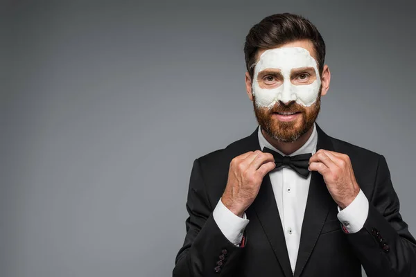 Happy and bearded man in suit with clay mask on face adjusting bow tie isolated on grey — Stock Photo