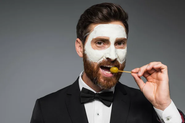 Bearded man in suit with clay mask on face holding toothpick and eating olive isolated on grey — Stock Photo