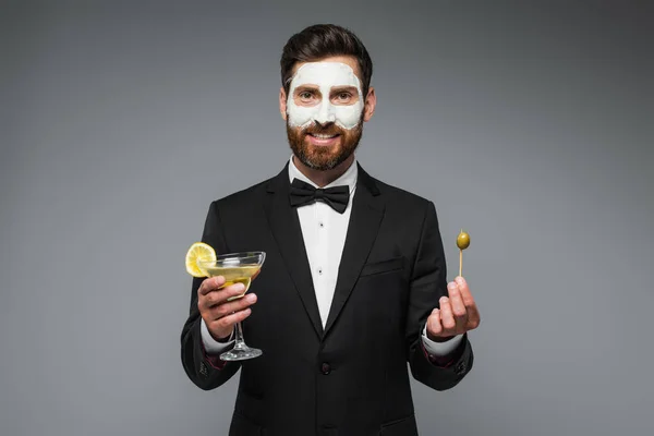 Happy man in suit with clay mask on face holding cocktail and toothpick with olive isolated on grey - foto de stock