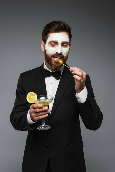 Бородатый мужчина в костюме с глиняной маской на лице, держа коктейль и зубочистку с оливкой, изолированной на сером — стоковое фото