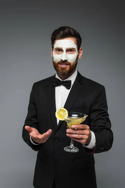Bearded man in tuxedo with clay mask on face holding cocktail and gesturing isolated on grey — Photo de stock