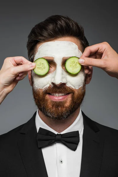 Women applying sliced cucumbers on eyes of bearded man in suit with clay mask  isolated on grey — Stock Photo