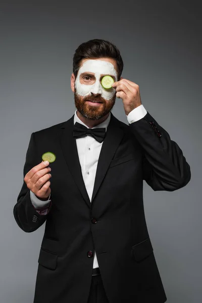 Bearded man in suit with clay mask on face holding slices of cucumber isolated on grey - foto de stock