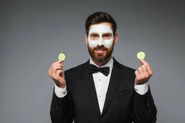 Happy bearded man in suit with clay mask on face holding slices of cucumber isolated on grey — Fotografia de Stock