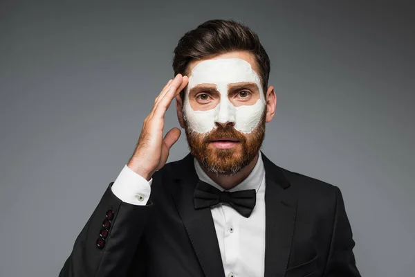 Bearded man in suit with clay mask on face adjusting hair isolated on grey — Stock Photo