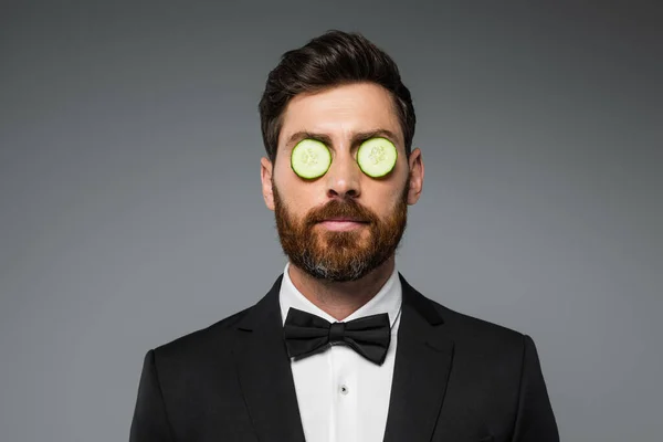 Bearded man in tuxedo with fresh sliced cucumber on eyes isolated on grey — Foto stock