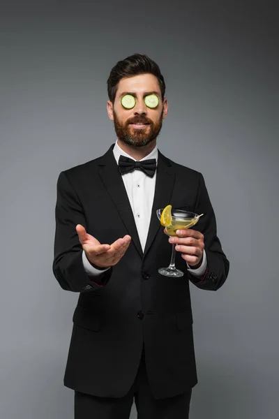 Bearded man in tuxedo with fresh sliced cucumber on eyes holding cocktail isolated on grey — Fotografia de Stock