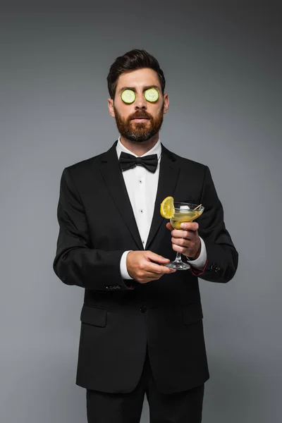 Bearded man in tuxedo with fresh sliced cucumber on eyes holding glass of cocktail isolated on grey - foto de stock