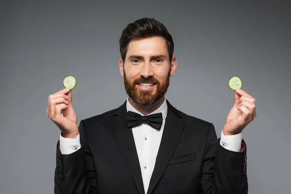 Bearded man in tuxedo holding fresh sliced cucumber and smiling isolated on grey — Stockfoto