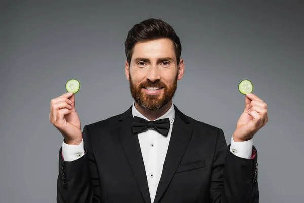 Bearded man in tuxedo holding fresh sliced cucumber and smiling isolated on grey — Stockfoto