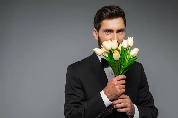 Bearded man in elegant tuxedo smelling fresh tulips isolated on grey — Stockfoto