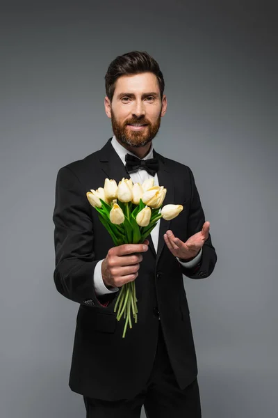 Bearded man in elegant suit with bow tie holding fresh tulips and smiling isolated on grey — Photo de stock