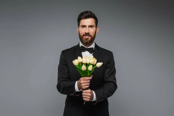 Бородатый мужчина в элегантном костюме с галстуком-бабочкой с букетом тюльпанов, изолированных на сером — стоковое фото
