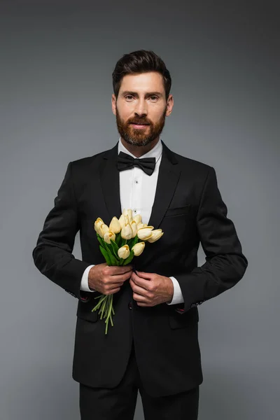Бородатый мужчина в элегантном смокинге с галстуком-бабочкой с букетом тюльпанов, изолированных на сером — стоковое фото