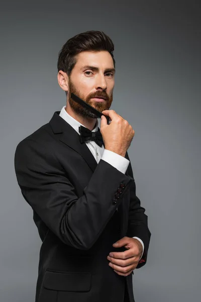 Man in elegant suit brushing beard with comb isolated on grey — Stock Photo