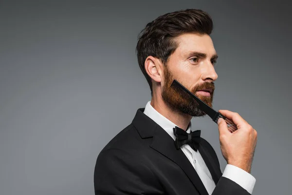 Man in tuxedo brushing beard with comb and looking away isolated on grey — Foto stock