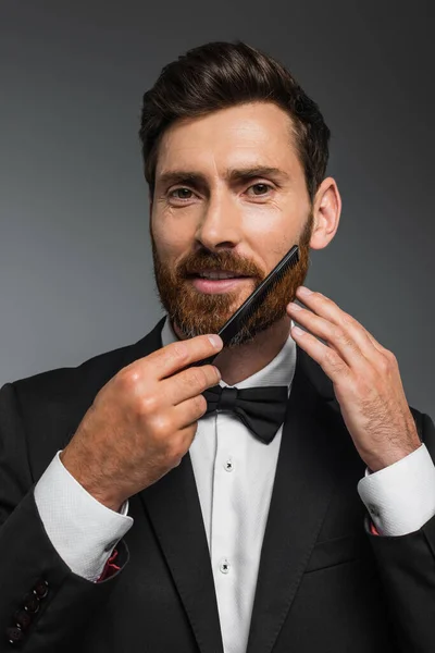 Portrait of happy man in tuxedo brushing beard with comb isolated on grey — Stock Photo