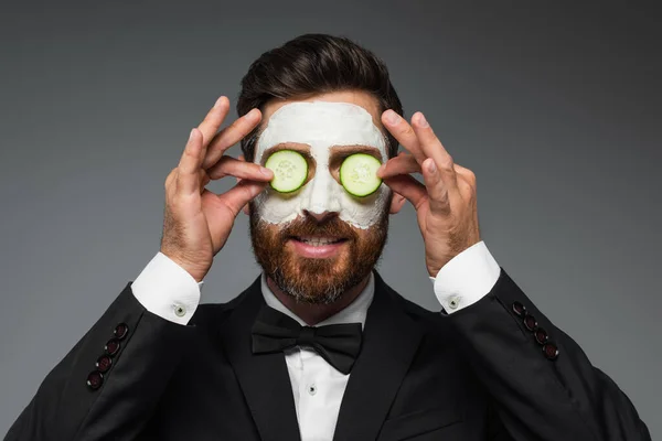 Бородатий чоловік з глиняною маскою на обличчі наносить нарізані огірки на очі ізольовані на сірому — стокове фото