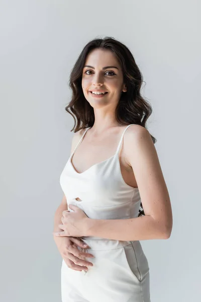 Portrait of smiling brunette woman in white clothes looking at camera isolated on grey - foto de stock