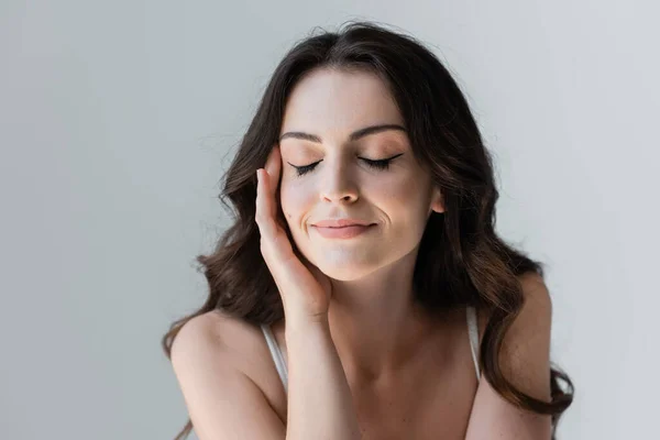 Smiling brunette woman closing eyes and touching face isolated on grey - foto de stock