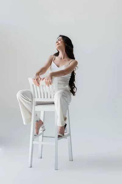 Full length of stylish brunette woman smiling while sitting on chair on grey background — Photo de stock