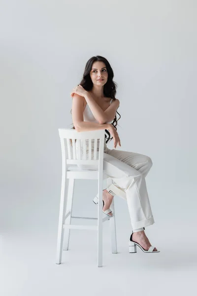 Full length of stylish woman touching shoulder while sitting on chair on grey background — Photo de stock