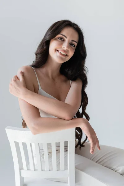 Positive long haired model in white clothes sitting on chair isolated on grey — Stock Photo