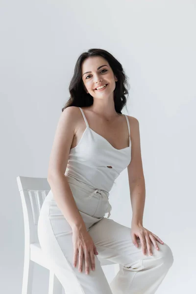 Pretty woman in white clothes smiling near chair isolated on grey — Stockfoto