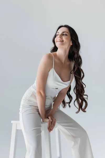 Positive long haired model sitting on chair and looking away isolated on grey — Foto stock