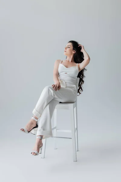 Full length of young model in white clothes and heels sitting on chair on grey background - foto de stock