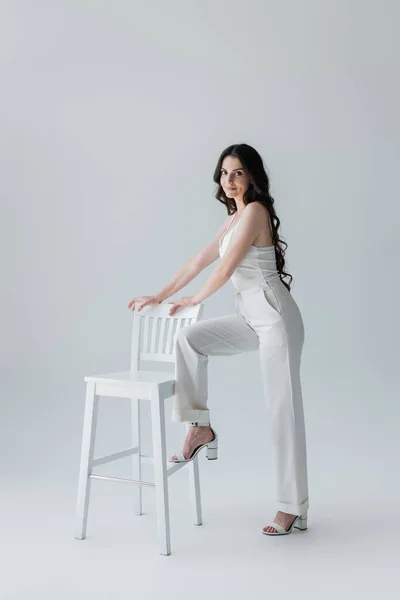 Smiling model in heels and white clothes looking at camera near chair on grey background — Stockfoto