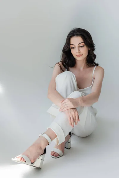 Brunette woman in heels sitting on grey background with sunlight — Photo de stock