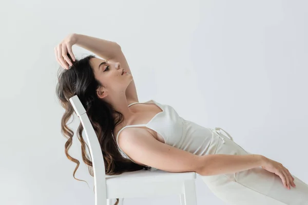 Side view of brunette woman touching hair near chair isolated on grey — Stockfoto