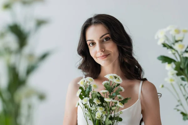 Smiling woman in white top looking at camera near blurred flowers isolated on grey — Stockfoto