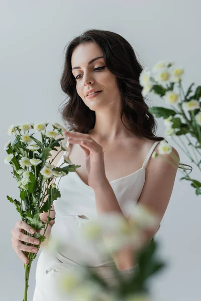 Pretty brunette woman in white top touching flowers isolated on grey — Stockfoto