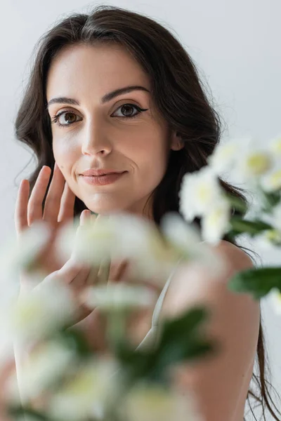 Portrait of brunette model with makeup looking at camera near blurred flowers isolated on grey — Stock Photo