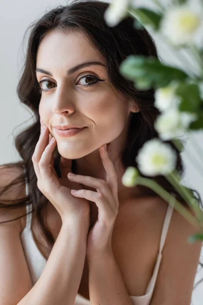 Brunette model with visage smiling at camera near blurred flowers isolated on grey — Fotografia de Stock