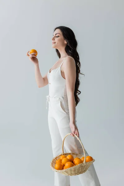 Smiling woman in white clothes holding basket with fresh oranges isolated on grey — Stock Photo