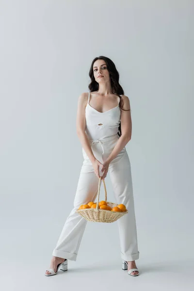 Full length of trendy woman holding basket with oranges on grey background — Stock Photo