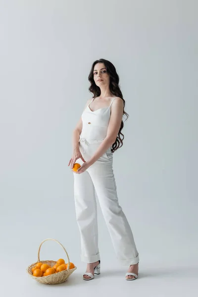 Stylish young woman holding orange near wicker basket on grey background — Foto stock