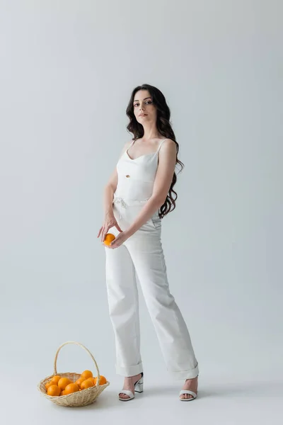 Stylish young woman holding orange near wicker basket on grey background — Stock Photo