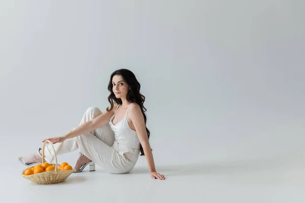 Pretty and stylish woman sitting near wicker basket with oranges on grey background — Stockfoto