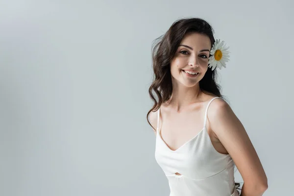 Happy brunette woman with chamomile in hair looking at camera isolated on grey — Stockfoto