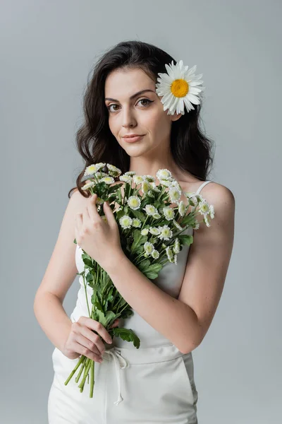 Pretty young woman holding bouquet of chamomiles while standing isolated on grey — Fotografia de Stock